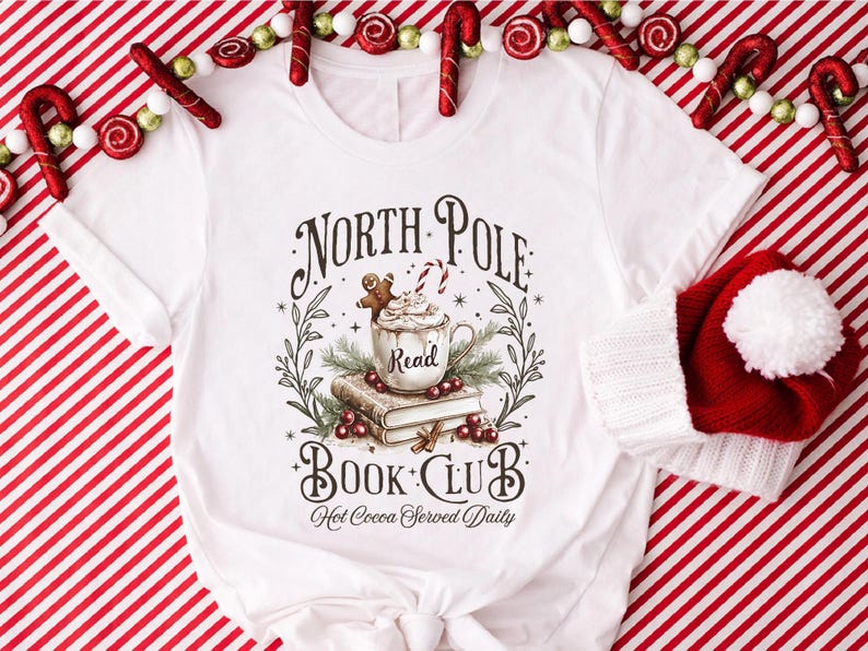 A festive T-shirt featuring a cozy Christmas-themed design that reads “North Pole Book Club Hot Cocoa Served Daily.” The artwork shows a cup of hot cocoa topped with whipped cream, a candy cane, and a gingerbread cookie resting on stacked books, surrounded by holly berries and pine branches; a perfect holiday shirt for book lovers and cozy winter readers who adore festive hot chocolate and Christmas vibes.
