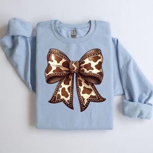 May include: A light blue sweatshirt featuring a large bow design. The bow has a brown and white cow print pattern with a detailed outline. The sweatshirt has long sleeves and a crew neck.
