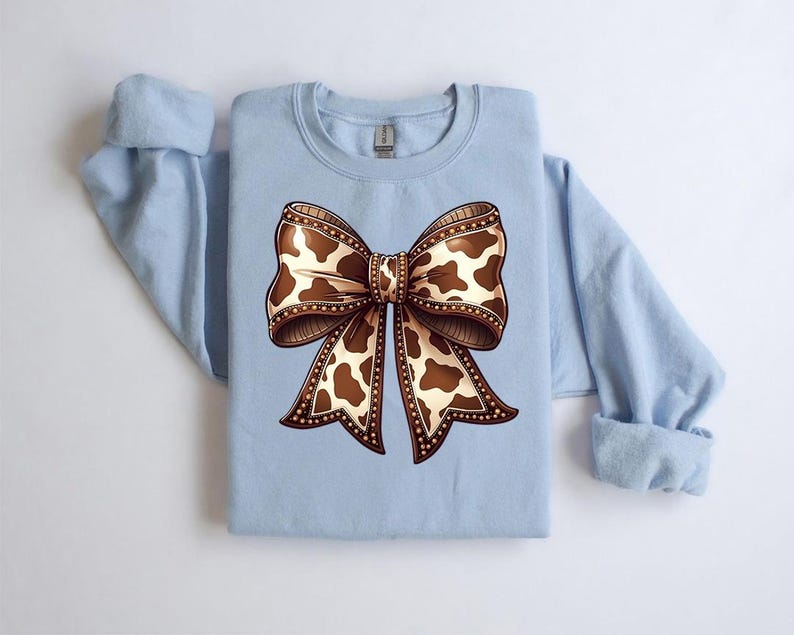 May include: A light blue sweatshirt featuring a large bow design. The bow has a brown and white cow print pattern with a detailed outline. The sweatshirt has long sleeves and a crew neck.