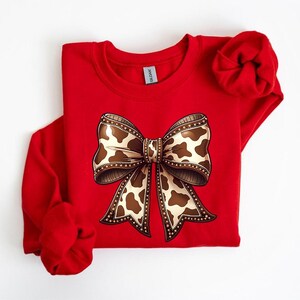 May include: A red sweatshirt featuring a large bow graphic. The bow has a brown and white cow print design with a decorative border. The sleeves are folded, and the Gildan tag is visible.
