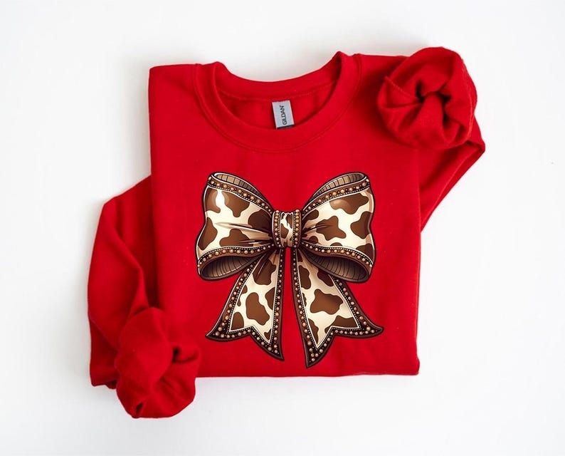 May include: A red sweatshirt featuring a large bow graphic. The bow has a brown and white cow print design with a decorative border. The sleeves are folded, and the Gildan tag is visible.