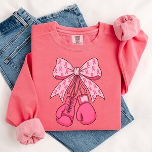 May include: A coral-colored sweatshirt featuring a graphic of a pink ribbon bow and boxing gloves, both adorned with pink ribbons. The sweatshirt has a crew neck and long sleeves with pink cuffs. The words "Comfort Colors" are on the tag.