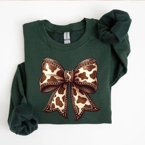 May include: A dark green sweatshirt featuring a large bow graphic. The bow has a brown and white cow print design with a detailed border. The sweatshirt is a solid color with a crew neck and long sleeves.