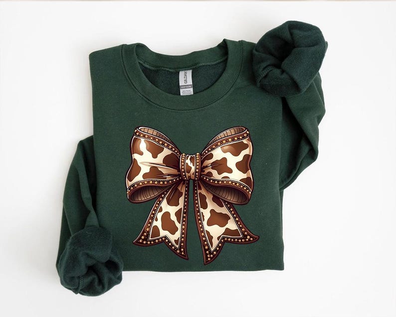May include: A dark green sweatshirt featuring a large bow graphic. The bow has a brown and white cow print design with a detailed border. The sweatshirt is a solid color with a crew neck and long sleeves.