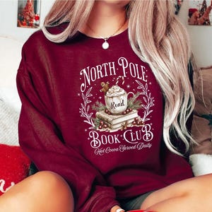 A festive T-shirt featuring a cozy Christmas-themed design that reads “North Pole Book Club Hot Cocoa Served Daily.” The artwork shows a cup of hot cocoa topped with whipped cream, a candy cane, and a gingerbread cookie resting on stacked books, surrounded by holly berries and pine branches; a perfect holiday shirt for book lovers and cozy winter readers who adore festive hot chocolate and Christmas vibes.