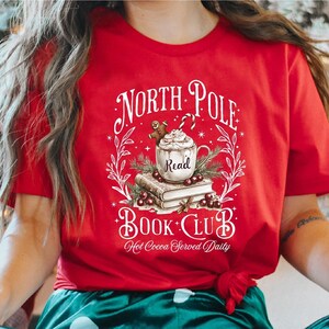 A festive T-shirt featuring a cozy Christmas-themed design that reads “North Pole Book Club Hot Cocoa Served Daily.” The artwork shows a cup of hot cocoa topped with whipped cream, a candy cane, and a gingerbread cookie resting on stacked books, surrounded by holly berries and pine branches; a perfect holiday shirt for book lovers and cozy winter readers who adore festive hot chocolate and Christmas vibes.