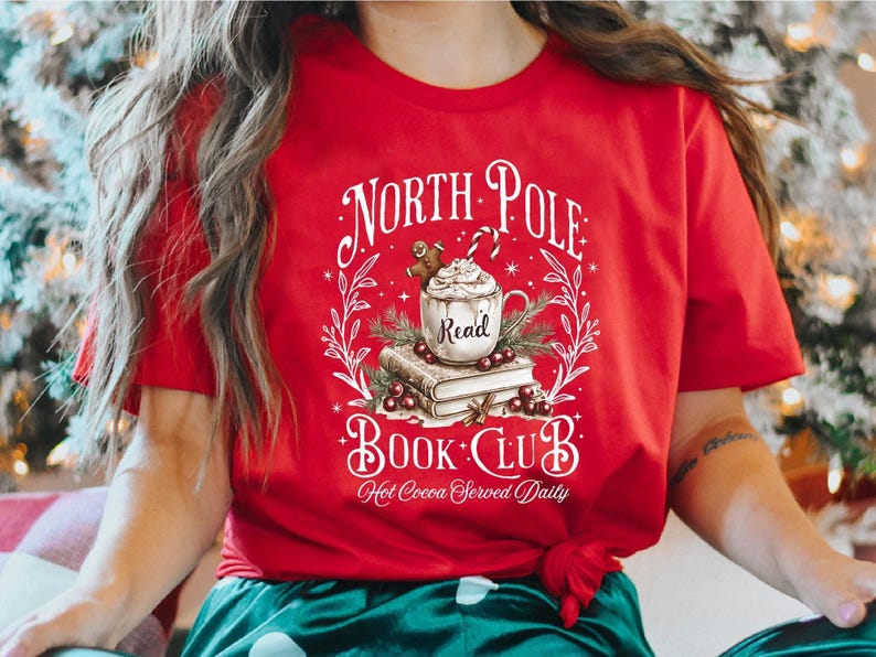 A festive T-shirt featuring a cozy Christmas-themed design that reads “North Pole Book Club Hot Cocoa Served Daily.” The artwork shows a cup of hot cocoa topped with whipped cream, a candy cane, and a gingerbread cookie resting on stacked books, surrounded by holly berries and pine branches; a perfect holiday shirt for book lovers and cozy winter readers who adore festive hot chocolate and Christmas vibes.