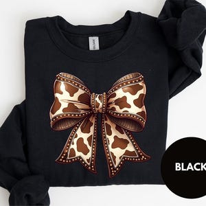 May include: Black sweatshirt featuring a large bow design with a cow print pattern in shades of brown and cream. The bow is detailed with a decorative border. The word "BLACK" is in a black circle.