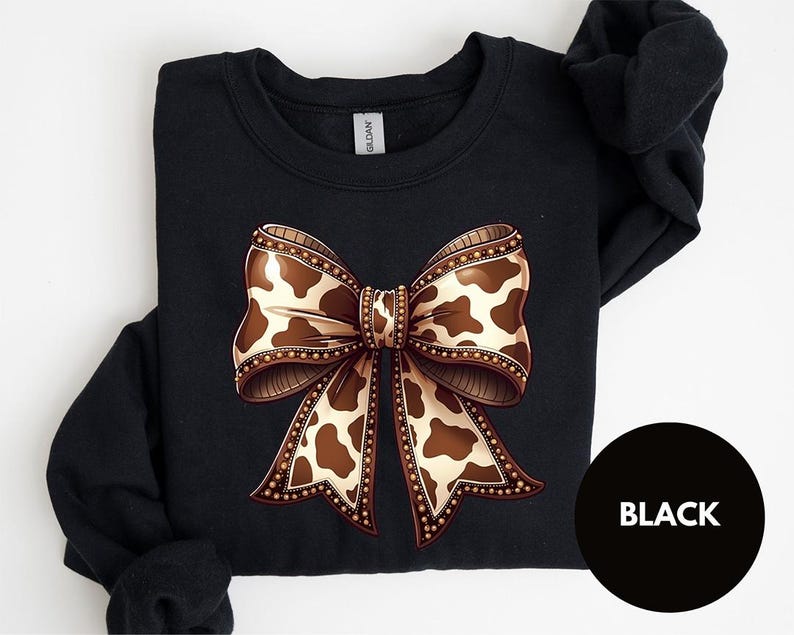 May include: Black sweatshirt featuring a large bow design with a cow print pattern in shades of brown and cream. The bow is detailed with a decorative border. The word "BLACK" is in a black circle.
