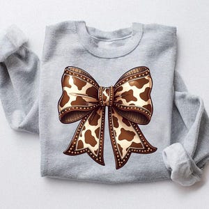 Graphic sweatshirt featuring an illustrated ribbon bow with cow print pattern inside the loops and tails; centered decorative bow design with rounded loops and flowing ribbon ends; western-inspired cow pattern with stitched border detailing; symmetrical layout across the front of the sweatshirt; clean illustrated artwork designed for casual everyday wear; professionally printed graphic with sharp outlines and clear pattern detail.