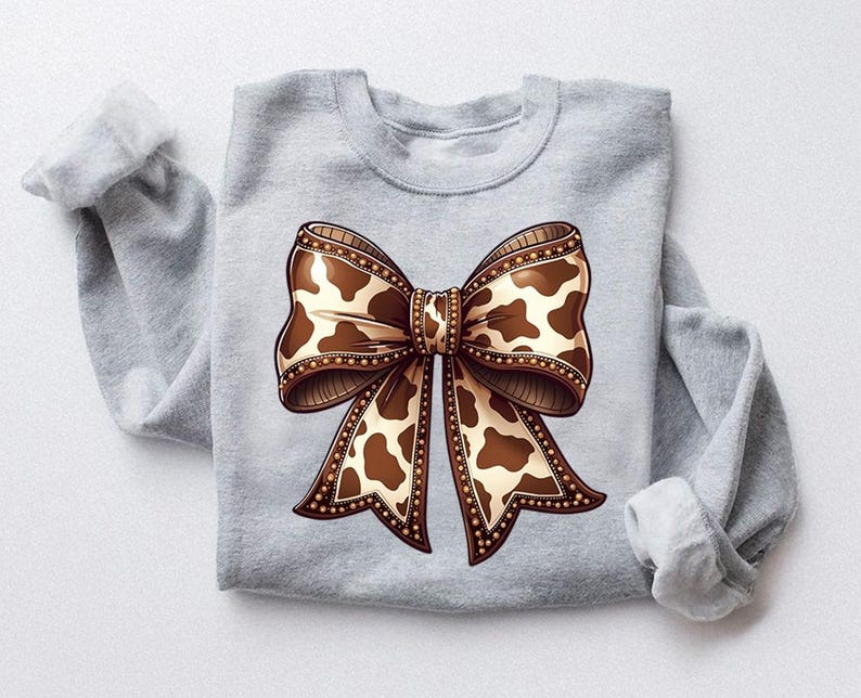 Graphic sweatshirt featuring an illustrated ribbon bow with cow print pattern inside the loops and tails; centered decorative bow design with rounded loops and flowing ribbon ends; western-inspired cow pattern with stitched border detailing; symmetrical layout across the front of the sweatshirt; clean illustrated artwork designed for casual everyday wear; professionally printed graphic with sharp outlines and clear pattern detail.