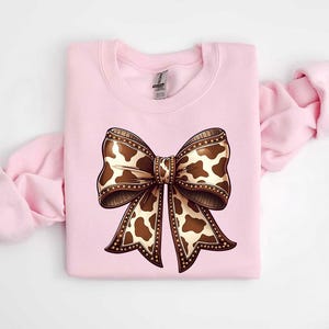 Graphic sweatshirt featuring an illustrated ribbon bow with cow print pattern inside the loops and tails; centered decorative bow design with rounded loops and flowing ribbon ends; western-inspired cow pattern with stitched border detailing; symmetrical layout across the front of the sweatshirt; clean illustrated artwork designed for casual everyday wear; professionally printed graphic with sharp outlines and clear pattern detail.