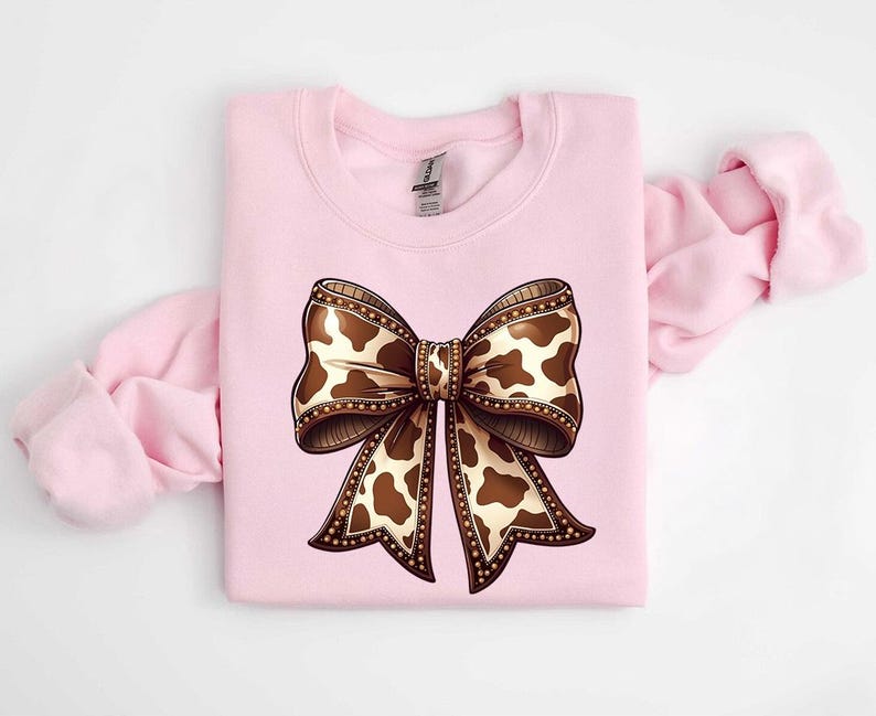 Graphic sweatshirt featuring an illustrated ribbon bow with cow print pattern inside the loops and tails; centered decorative bow design with rounded loops and flowing ribbon ends; western-inspired cow pattern with stitched border detailing; symmetrical layout across the front of the sweatshirt; clean illustrated artwork designed for casual everyday wear; professionally printed graphic with sharp outlines and clear pattern detail.