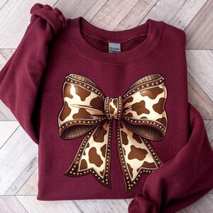 May include: A maroon sweatshirt featuring a large bow design. The bow has a cowhide pattern in shades of brown and cream, outlined with a beaded border. The sweatshirt is folded on a light wood-grain surface.