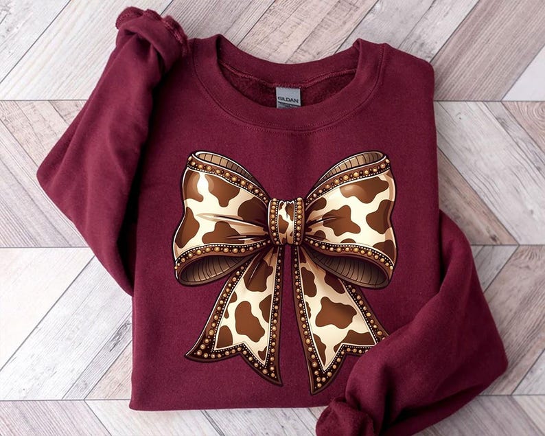 May include: A maroon sweatshirt featuring a large bow design. The bow has a cowhide pattern in shades of brown and cream, outlined with a beaded border. The sweatshirt is folded on a light wood-grain surface.