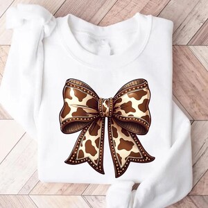 Graphic sweatshirt featuring an illustrated ribbon bow with cow print pattern inside the loops and tails; centered decorative bow design with rounded loops and flowing ribbon ends; western-inspired cow pattern with stitched border detailing; symmetrical layout across the front of the sweatshirt; clean illustrated artwork designed for casual everyday wear; professionally printed graphic with sharp outlines and clear pattern detail.