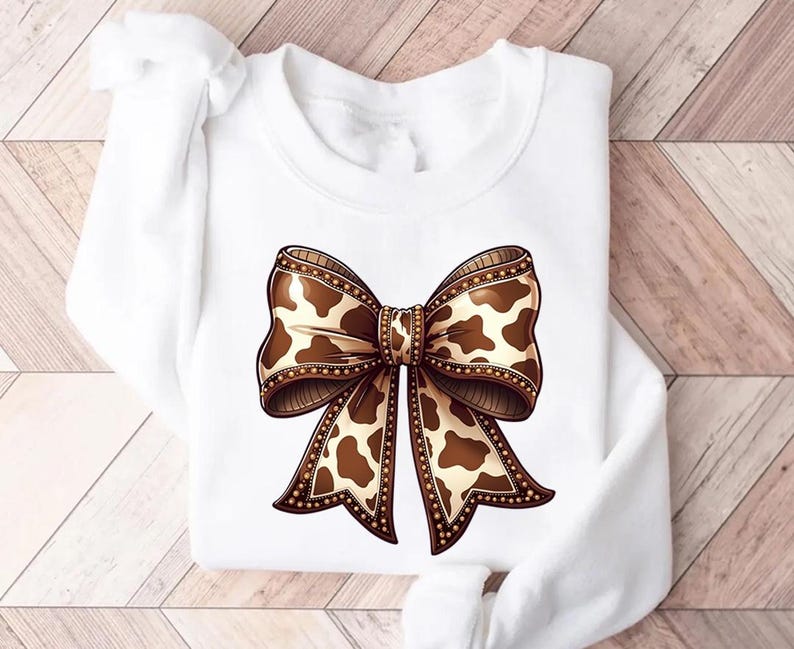 Graphic sweatshirt featuring an illustrated ribbon bow with cow print pattern inside the loops and tails; centered decorative bow design with rounded loops and flowing ribbon ends; western-inspired cow pattern with stitched border detailing; symmetrical layout across the front of the sweatshirt; clean illustrated artwork designed for casual everyday wear; professionally printed graphic with sharp outlines and clear pattern detail.