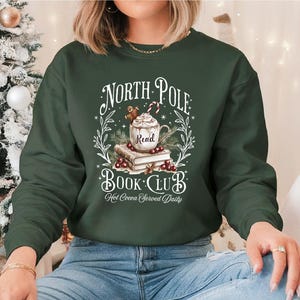 A festive T-shirt featuring a cozy Christmas-themed design that reads “North Pole Book Club Hot Cocoa Served Daily.” The artwork shows a cup of hot cocoa topped with whipped cream, a candy cane, and a gingerbread cookie resting on stacked books, surrounded by holly berries and pine branches; a perfect holiday shirt for book lovers and cozy winter readers who adore festive hot chocolate and Christmas vibes.