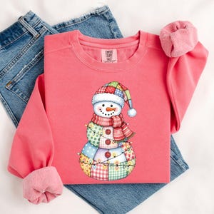 May include: A coral-colored sweatshirt featuring a snowman graphic. The snowman wears a patchwork hat, scarf, and is decorated with string lights. The sweatshirt is displayed on top of blue jeans.