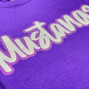 May include: A purple t-shirt with the word "Mustangs" in a silver and glittery purple design. The text is in a cursive font and is centered on the shirt.