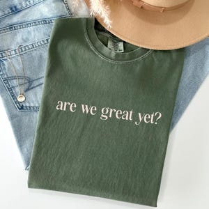May include: An olive green t-shirt with the phrase "are we great yet?" in white lettering. The shirt is laid on top of light blue denim jeans, with a gold necklace and a tan fedora hat. A casual, everyday outfit.