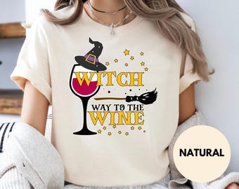May include: A natural-colored t-shirt featuring a graphic of a wine glass wearing a witch hat, a broomstick, and yellow stars. The text reads WITCH WAY TO THE WINE in yellow block letters. The shirt has short sleeves and a crew neckline.