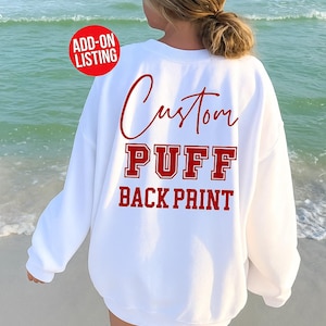 Könnte beinhalten: Weißes Sweatshirt mit rotem Aufdruck "Custom PUFF BACK PRINT". Das Sweatshirt wird von hinten gezeigt, mit dem Meer im Hintergrund. Ein "ADD-ON LISTING"-Aufkleber befindet sich in der oberen linken Ecke.