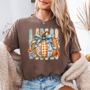 Peut inclure: Un t-shirt marron à manches courtes avec un motif central représentant une citrouille ornée d'un nœud bleu. La citrouille présente des sections à motifs léopard, vichy orange, floraux et à pois sur un fond de rayures verticales bleu sarcelle.