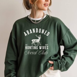 A sweatshirt and shirt featuring the Abandoned Hunting Wives Social Club design with a vintage deer illustration and bold rustic lettering, perfect for hunting wives, fall season wear, hunting camp gatherings, and cozy casual outfits.