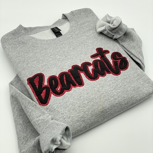 May include: A heather gray sweatshirt with the word "Bearcats" in a black, glitter-outlined font across the chest. The sweatshirt has a crew neck and long sleeves with ribbed cuffs. The garment is folded on a white surface.