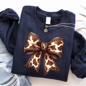 May include: Navy blue sweatshirt featuring a large bow design with a cowhide pattern in shades of brown and cream. The bow is embellished with a decorative border. A silver necklace with a pendant rests on the sweatshirt.