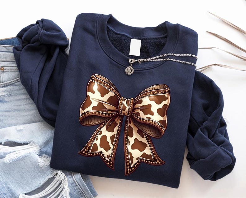 May include: Navy blue sweatshirt featuring a large bow design with a cowhide pattern in shades of brown and cream. The bow is embellished with a decorative border. A silver necklace with a pendant rests on the sweatshirt.