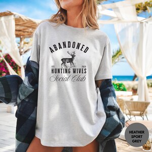 A sweatshirt and shirt featuring the Abandoned Hunting Wives Social Club design with a vintage deer illustration and bold rustic lettering, perfect for hunting wives, fall season wear, hunting camp gatherings, and cozy casual outfits.