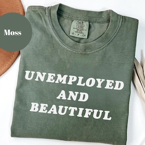 May include: A moss green t-shirt with the words "UNEMPLOYED AND BEAUTIFUL" printed in white. The shirt has a crew neck and short sleeves. A brown hat and dried plant are in the background.