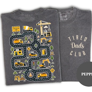 May include: Two gray t-shirts. One features a colorful construction zone design with trucks, a crane, and road details. The other shirt has the text "Tired Dads Club" in a cream color. The shirts are a pepper color.