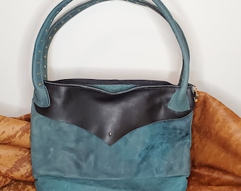 Op de afbeelding: Een tealblauwe leren handtas met een zwarte leren klep en een dubbele band. De tas heeft een gestructureerd oppervlak en staat op een bruin oppervlak.
