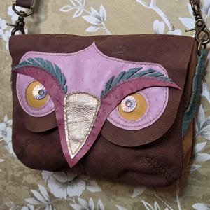May include: A brown leather crossbody bag with an owl design. The owl features pink, brown, and gold accents, with green feather details. The bag has a brown leather strap and metal hardware.