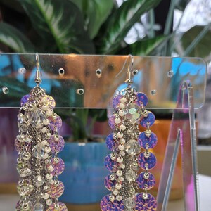 May include: A pair of long silver dangle earrings with iridescent purple and white round sequins and clear beads.
