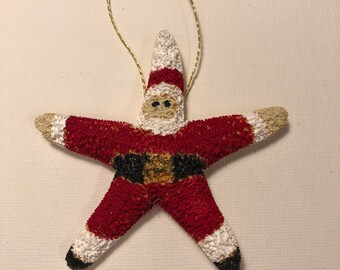 May include: A red and white Santa Claus ornament shaped like a starfish with a black belt and white trim. The ornament has a gold string for hanging.