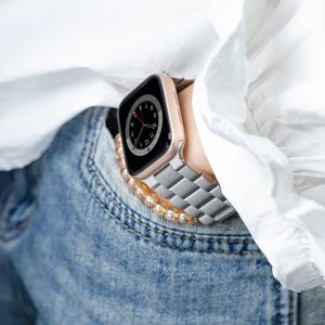 Op de afbeelding: Een roségouden Apple Watch met een zilveren metalen band. Het horloge wordt gedragen aan de pols van een persoon en is gedeeltelijk bedekt door een wit shirt en een blauwe jeans.