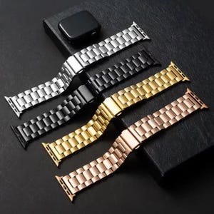 May include: Four metal watch straps in silver, black, gold, and rose gold. Each strap is made of rectangular links and has a clasp. A black watch is visible in the background.