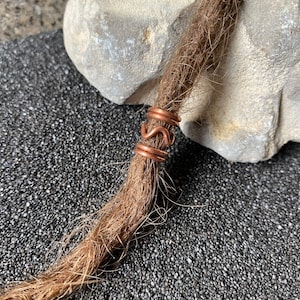 Può includere: Un perno per dreadlock in filo di rame con un design a spirale decorativo. Il perno è avvolto attorno a una dreadlock marrone.