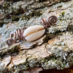 Puede incluir: Una concha de cauri blanca está envuelta en alambre de cobre, creando un accesorio para el cabello decorativo. El alambre está retorcido en forma de espiral.