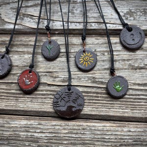 Op de afbeelding: Een verzameling handgemaakte keramische hangerkettingen. Elke hanger is een donkergrijze of rode schijf met unieke reliëfontwerpen, waaronder een zon, een hand en andere symbolen. De hangers hangen aan zwarte koorden.