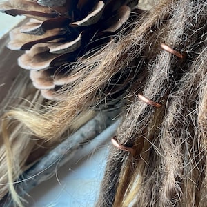 Könnte beinhalten: Drei Kupferhaarreifen werden verwendet, um Dreadlocks zu fixieren. Die Ringe sind glänzend kupferfarben und liegen eng beieinander auf den Dreadlocks.