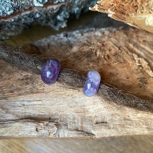 Puede incluir: Dos pequeñas piedras de amatista púrpura, cada una con una forma redonda y lisa, se muestran sobre una pieza de madera marrón.