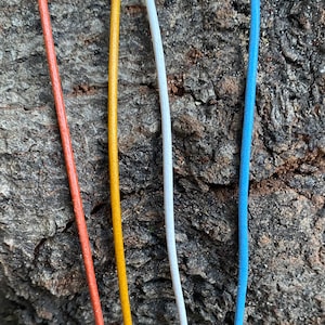 Puede incluir: Cuatro cordones de diferentes colores: naranja, amarillo, blanco y azul. Los cordones est&aacute;n colocados sobre una superficie de madera rugosa y marr&oacute;n.