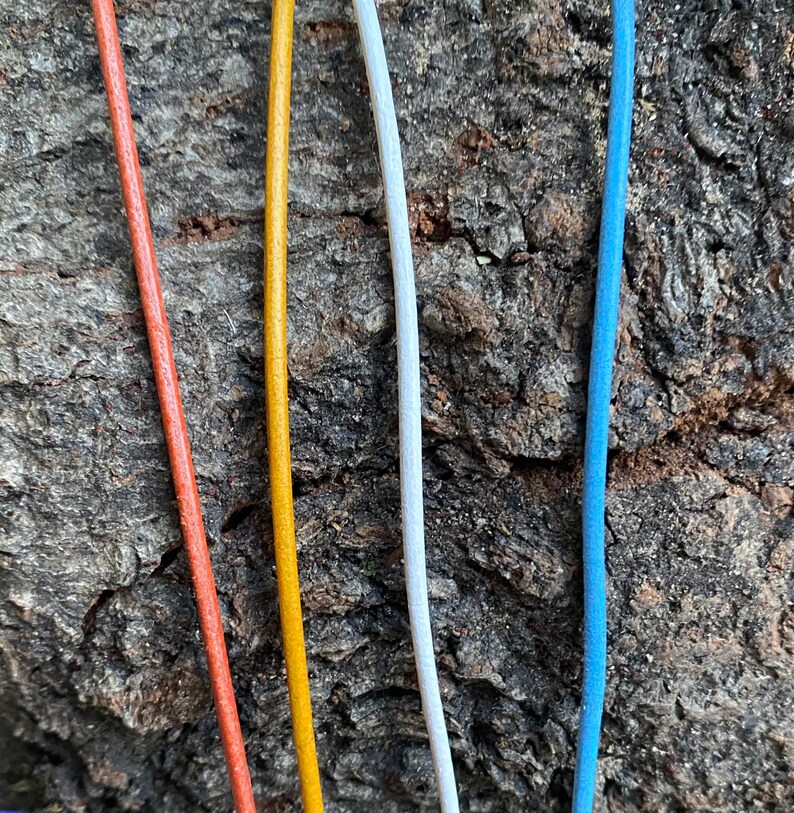 Puede incluir: Cuatro cordones de diferentes colores: naranja, amarillo, blanco y azul. Los cordones est&aacute;n colocados sobre una superficie de madera rugosa y marr&oacute;n.