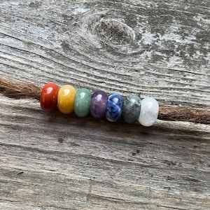 Könnte beinhalten: Ein Satz von sieben Chakra-Perlen in einem Regenbogen-Farbschema, auf einer braunen Schnur aufgefädelt. Die Perlen bestehen aus verschiedenen Edelsteinen, darunter roter Jaspis, gelber Citrin, grüner Aventurin, Amethyst, Lapislazuli, grauer Labradorit und klarer Quarz.