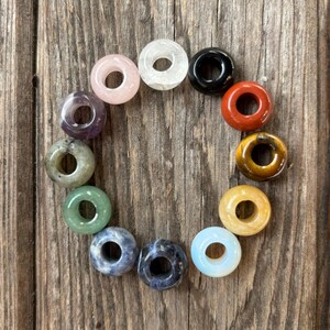 Puede incluir: Un conjunto de 12 cuentas de piedras preciosas en forma de rosquilla en varios colores, incluyendo transparente, rosa, negro, rojo, morado, verde, marrón, amarillo, azul y blanco. Las cuentas están dispuestas en círculo sobre una superficie de madera.